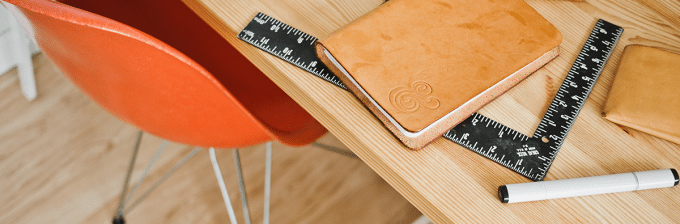 a top down image of a desktop with a leather bound notebook next to a square ruler and a pen