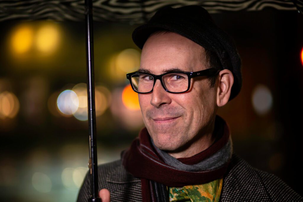 Keith Lowell Jensen Smiling At The Camera Wearing Black Glasses Under An Umbrella Outside In The Rain While He Is Wearing A Scarf And A Winter Coat